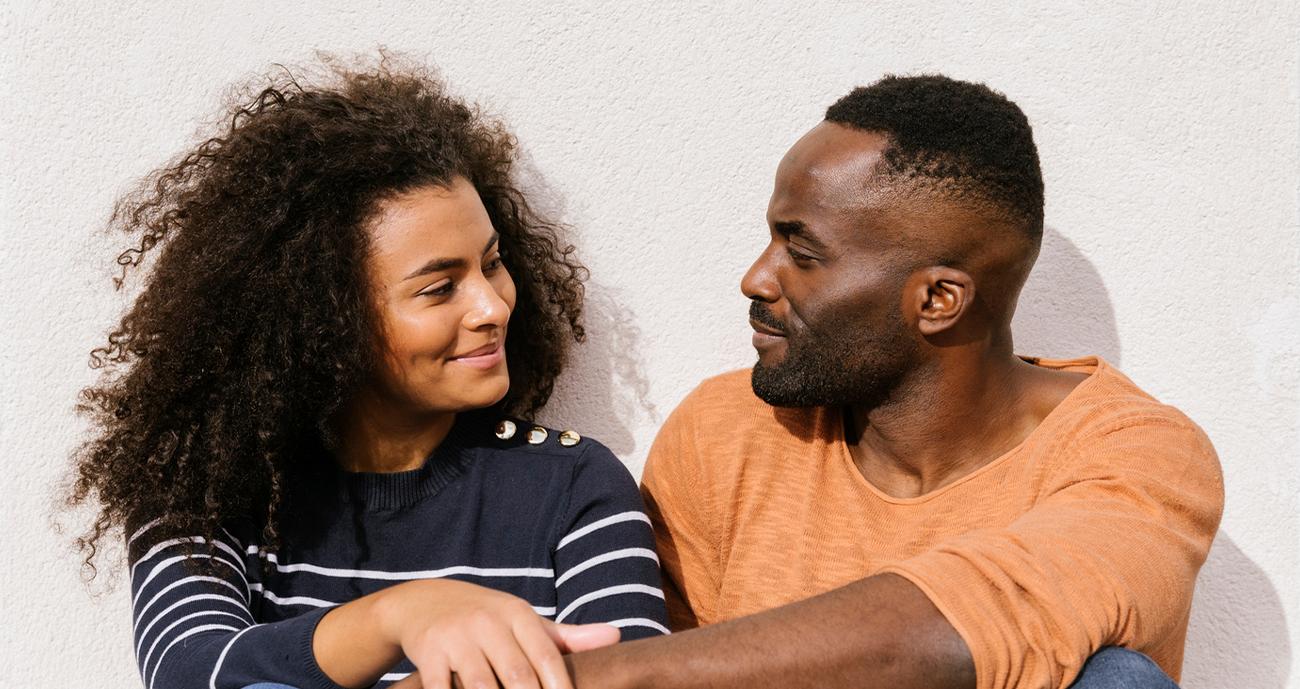 A couple sitting together staring into each other's eyes. The man is wearing an orange sweatshirt and the woman is wearing a stripped top.