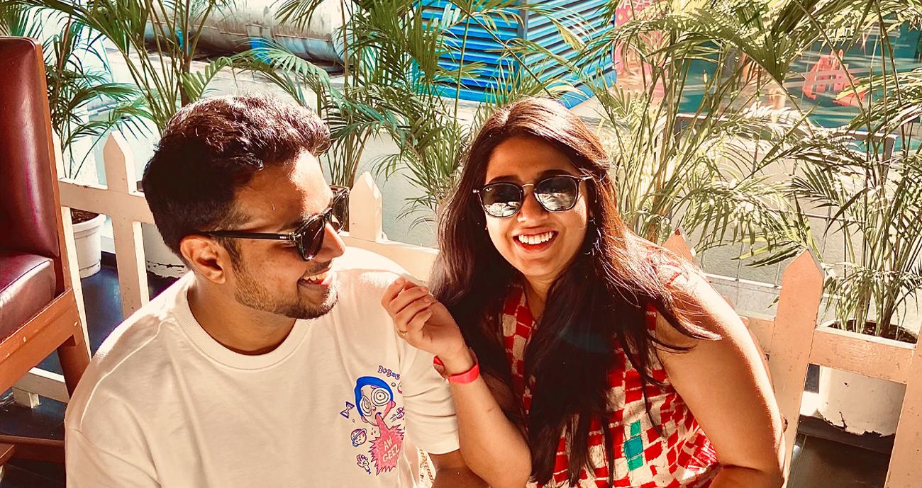Deepti and Kishan smiling at the camera while out at a restaurant together. Kishan is wearing a white T-shirt and Deepti is wearing a bright patterned blouse. They are sat in front of greenery.