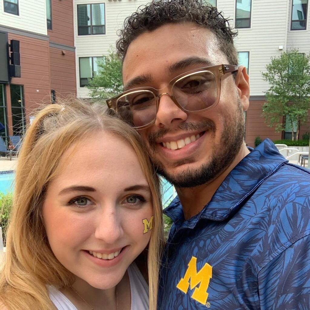 Jess and Nick are standing together and smiling at the camera. Jess is wearing a white tank top and Nick is wearing a blue print shirt.
