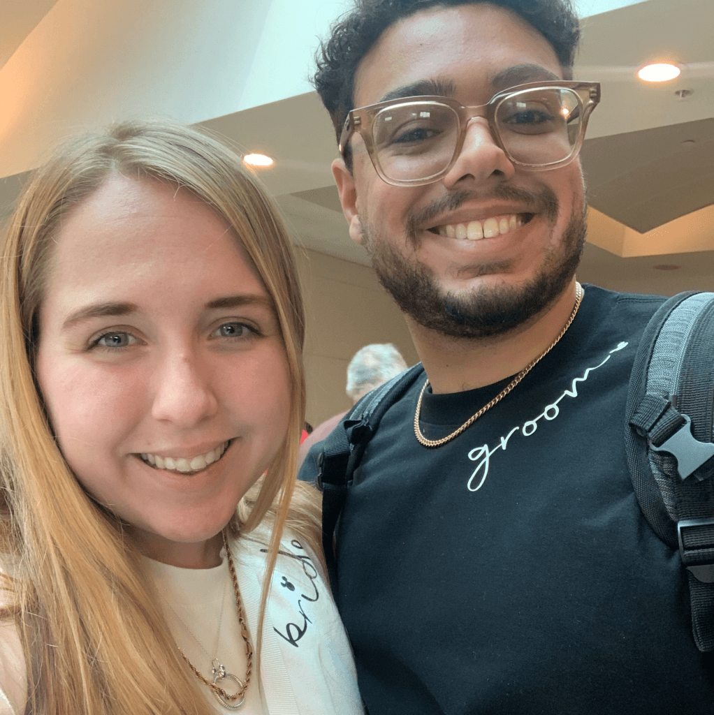 Jess and Nick are standing together smiling at the camera. Nick is wearing a black T-shirt with the word 'groom' on the front of it. Jess is wearing a white t-shirt with the word 'bride'.