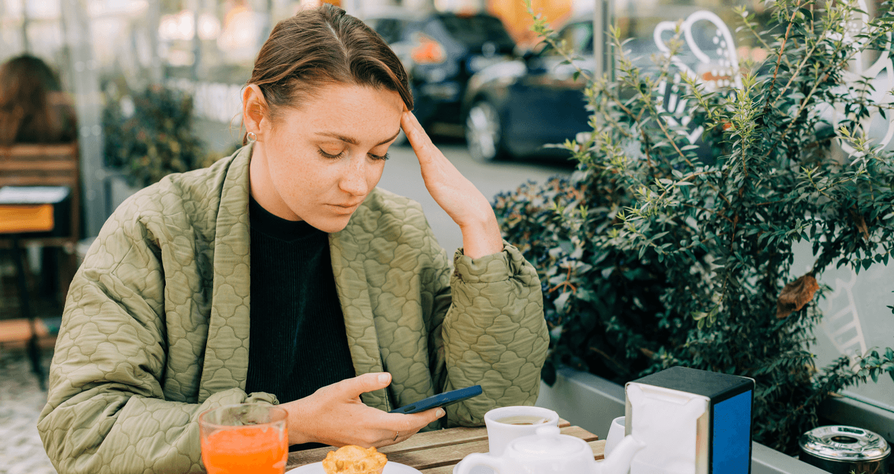 Young woman looking stressed and reflective while holding her phone at an outdoor cafe, illustrating the confusion and emotional toll that love bombing can bring in relationships.