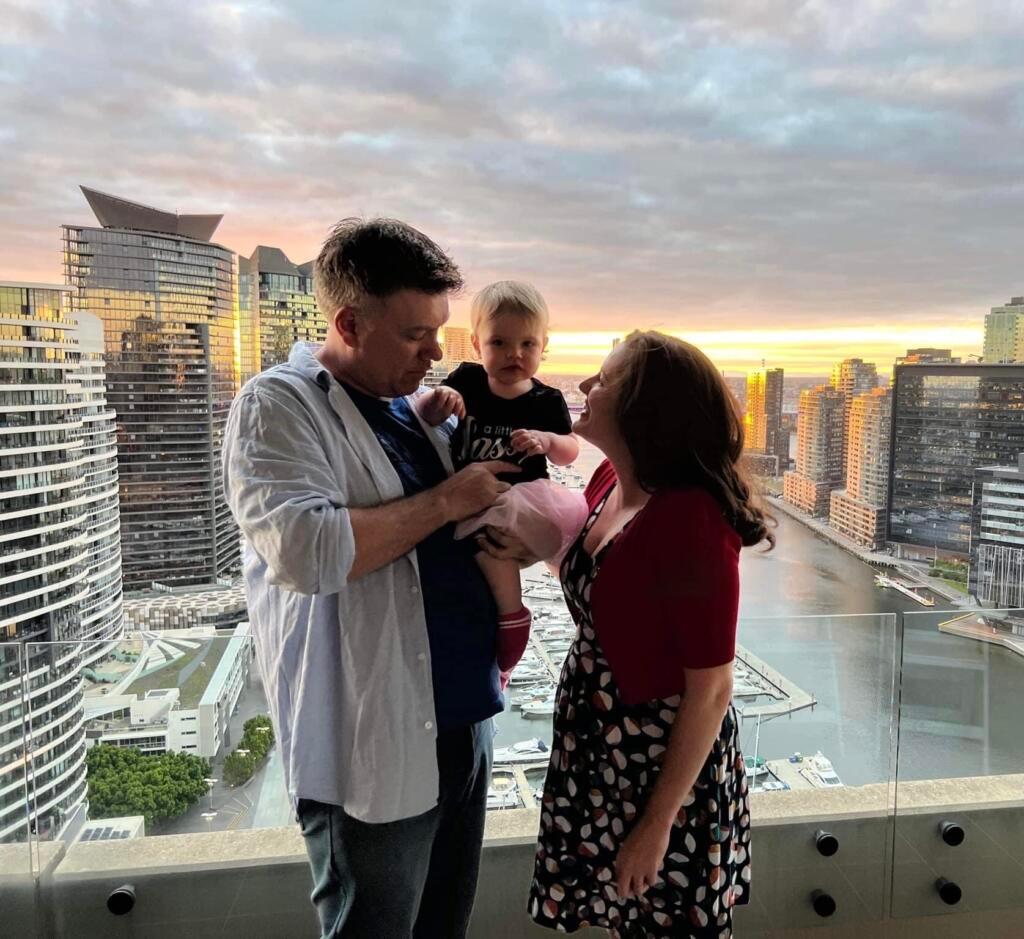 Jude and Ben are standing together. Ben is holding their daughter in his arms.